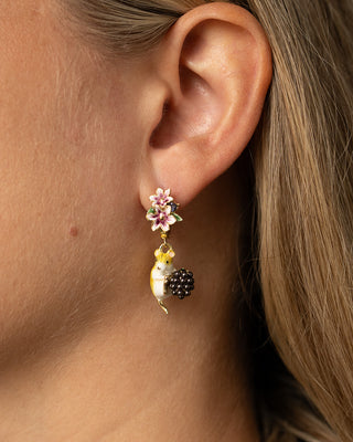 A close-up of a woman's ear wearing a dangling earring. The earring features a pink and white flower with green leaves at the top, hanging from a gold-toned post, with a small yellow and white mouse charm and a black berry charm below.
