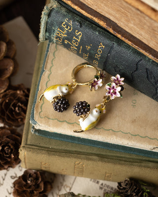 A pair of gold-tone hoop earrings with white and yellow mouse charms, black berry clusters, and pink-and-white flowers, displayed on vintage books with pinecones around them.