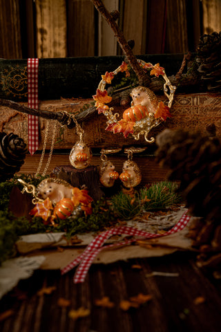 Autumn-themed jewelry set featuring small hedgehog pendants and matching earrings decorated with orange pumpkins, gold-toned leaves, and red-and-white gingham ribbon accents, styled on rustic branches and moss.