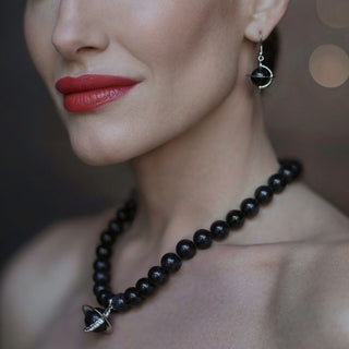 Close-up of a woman wearing a dark blue sandstone beaded necklace with an orb pendant and matching fishhook orb earrings with a blurred background.