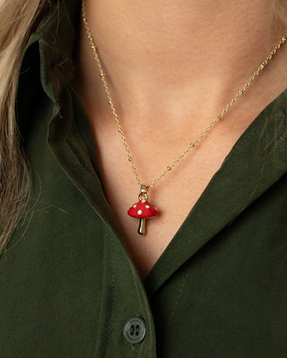 A close-up of a person wearing a delicate gold chain necklace with a small red mushroom toadstool pendant with white spots, styled with an olive green shirt.